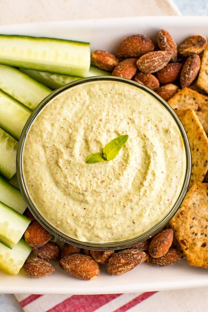 A pesto de amêndoa mergulha numa tigela num tabuleiro rodeado de amêndoas, bolachas e fatias de pepino.