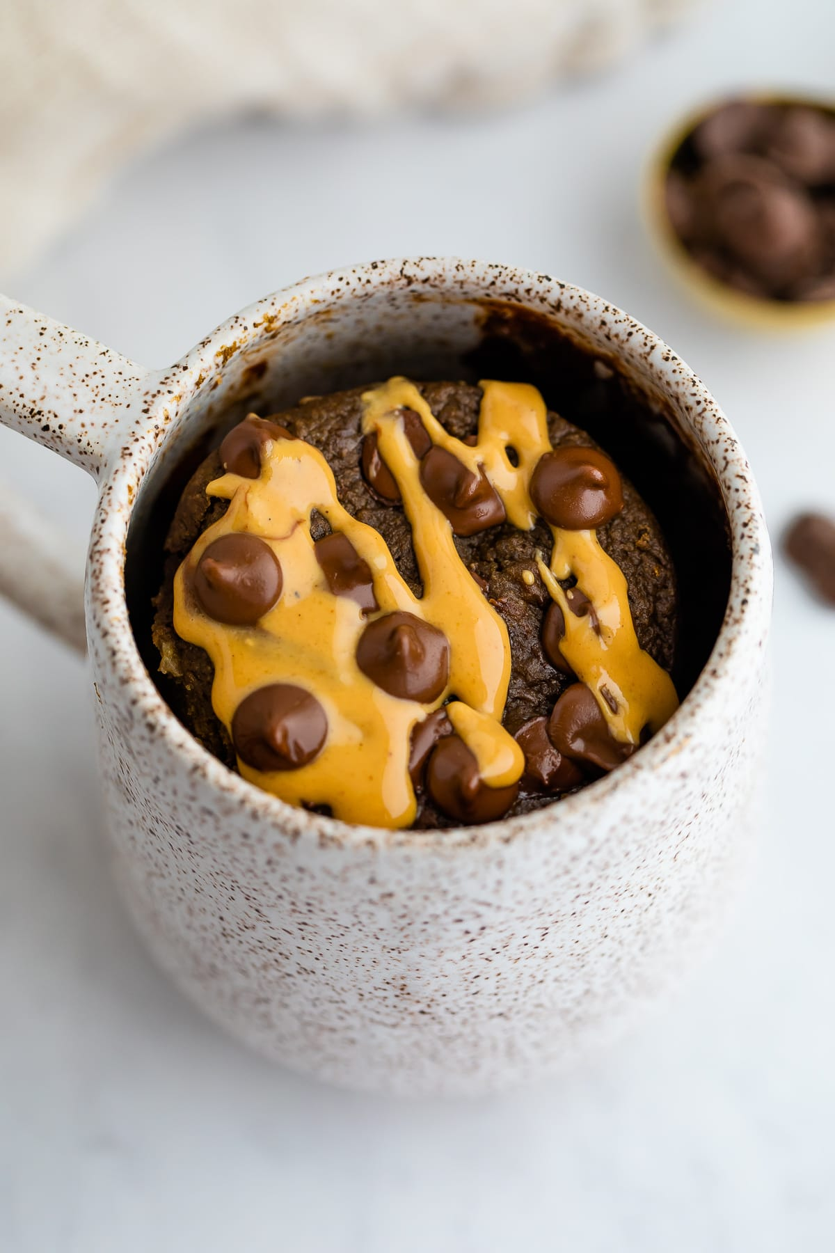 Bolo de caneca de proteína de chocolate, regado com manteiga de amendoim e coberto com pedaços de chocolate.