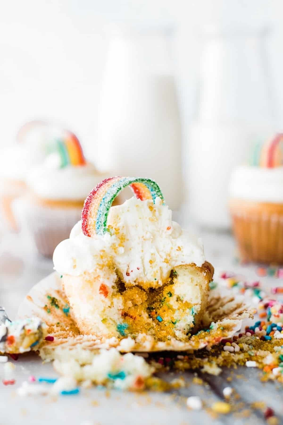 cupcake de funfetti com cobertura branca e rebuçados azedos no topo em forma de arco-íris, salpicos caindo do centro