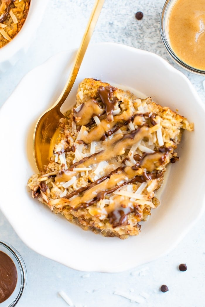 Uma fatia de papa de aveia assada em uma tigela com uma colher de ouro. A aveia é coberta com flocos de coco e chuviscos de caramelo e chocolate.