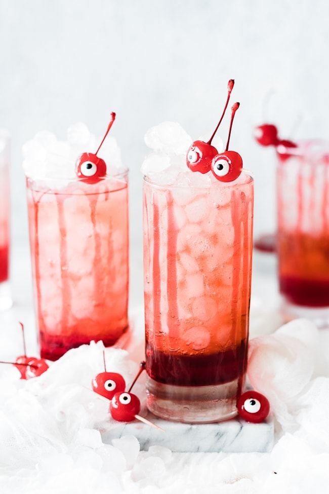 duas chávenas cheias de gelo, bebida rosa com xarope vermelho no fundo e dois globos oculares de cereja