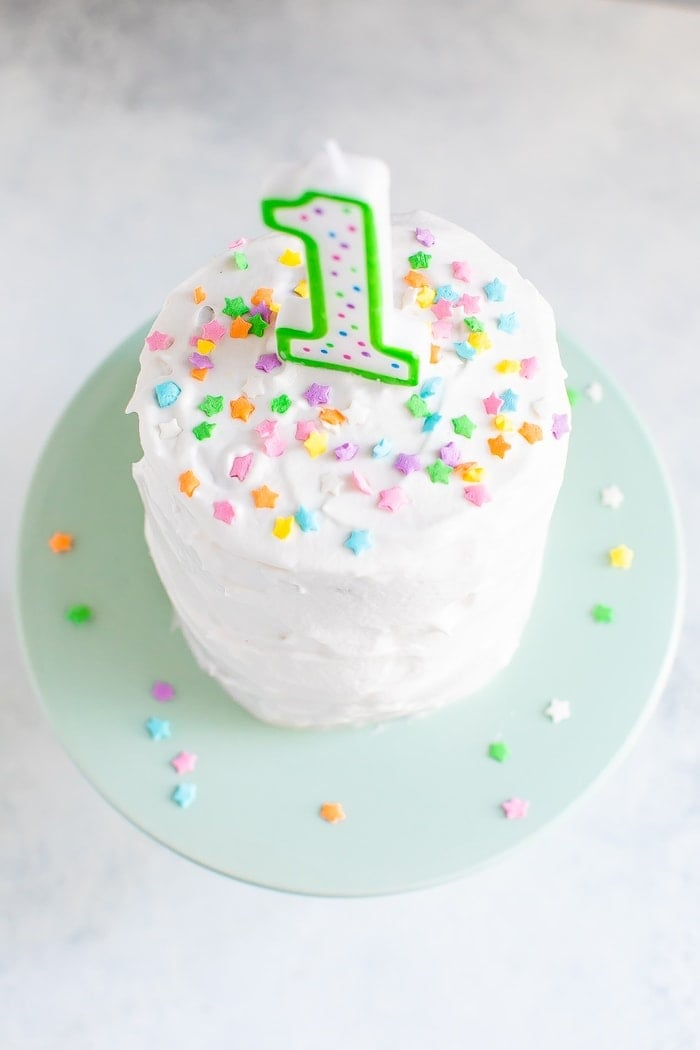 Pequeno bolo esmagador com cobertura branca, uma vela de aniversário de "1" e salpicos de estrelas coloridas.