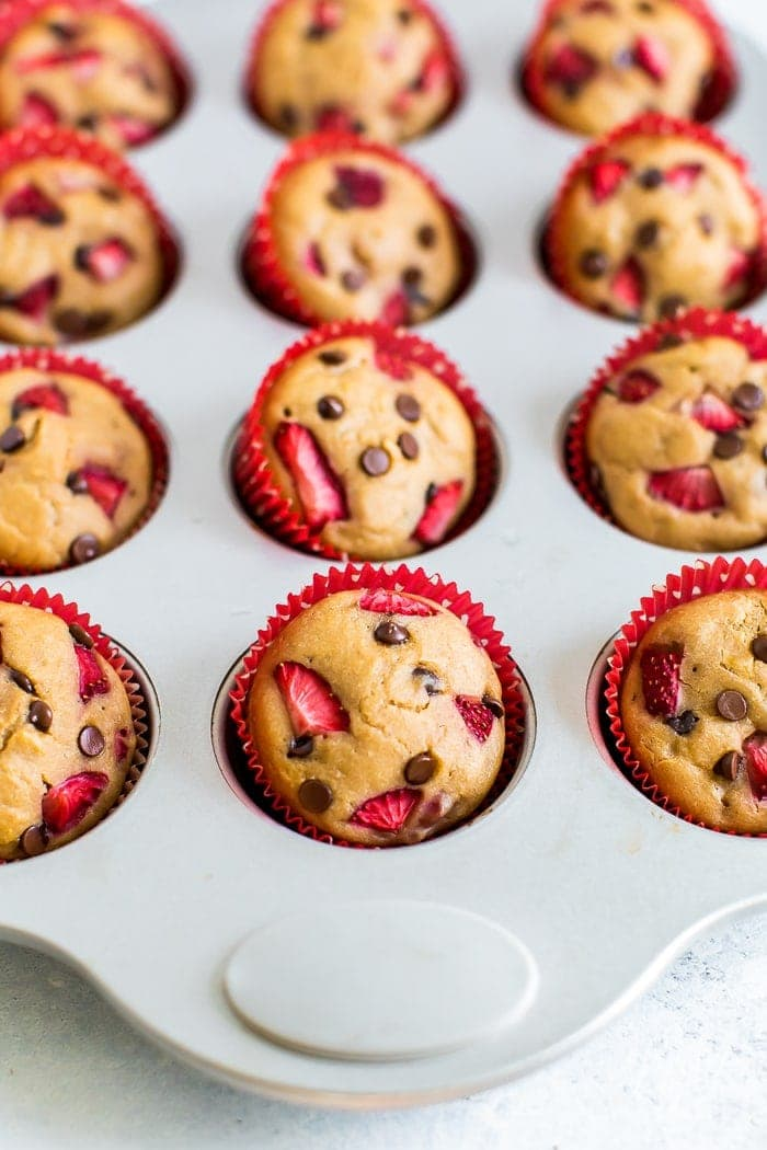 Muffins de proteína de chocolate de morango em bolinhos vermelhos numa lata de muffin. Os muffins são cobertos com morangos picados e mini pedaços de chocolate.