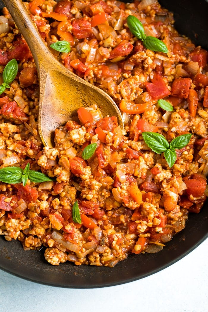 Colher de madeira mexendo uma panela de molho tempeh bolognese que é coberta com manjericão fresco.