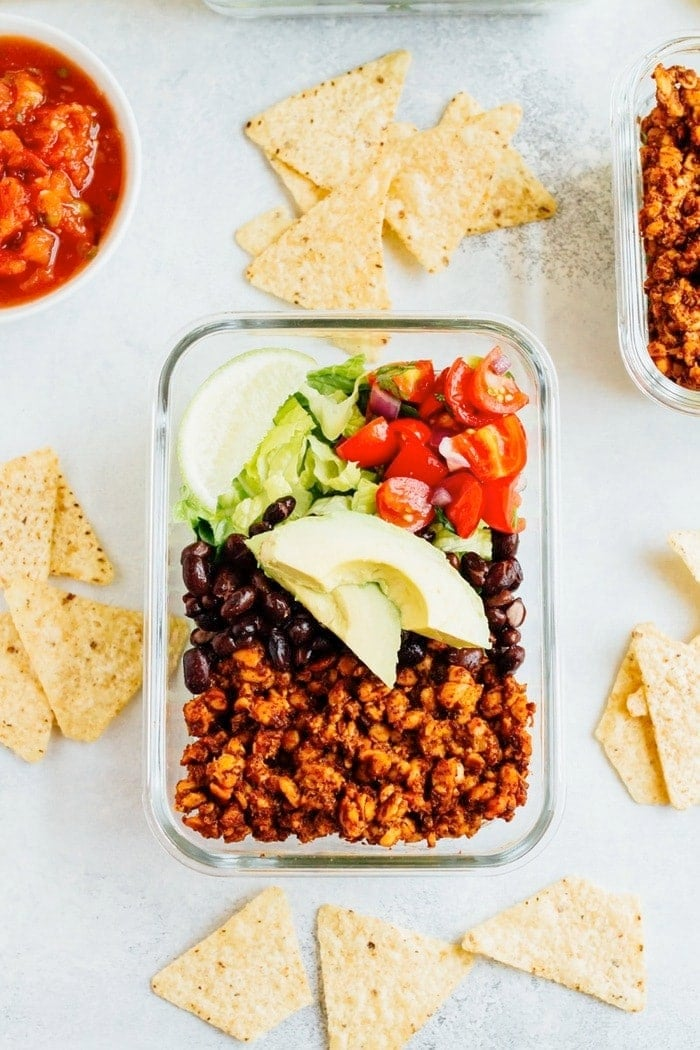 Um prato de vidro com salada de tempeh, incluindo tempeh, feijão preto, tomate alface e lima. Salsa, tempeh, e batatas fritas na mesa ao lado.