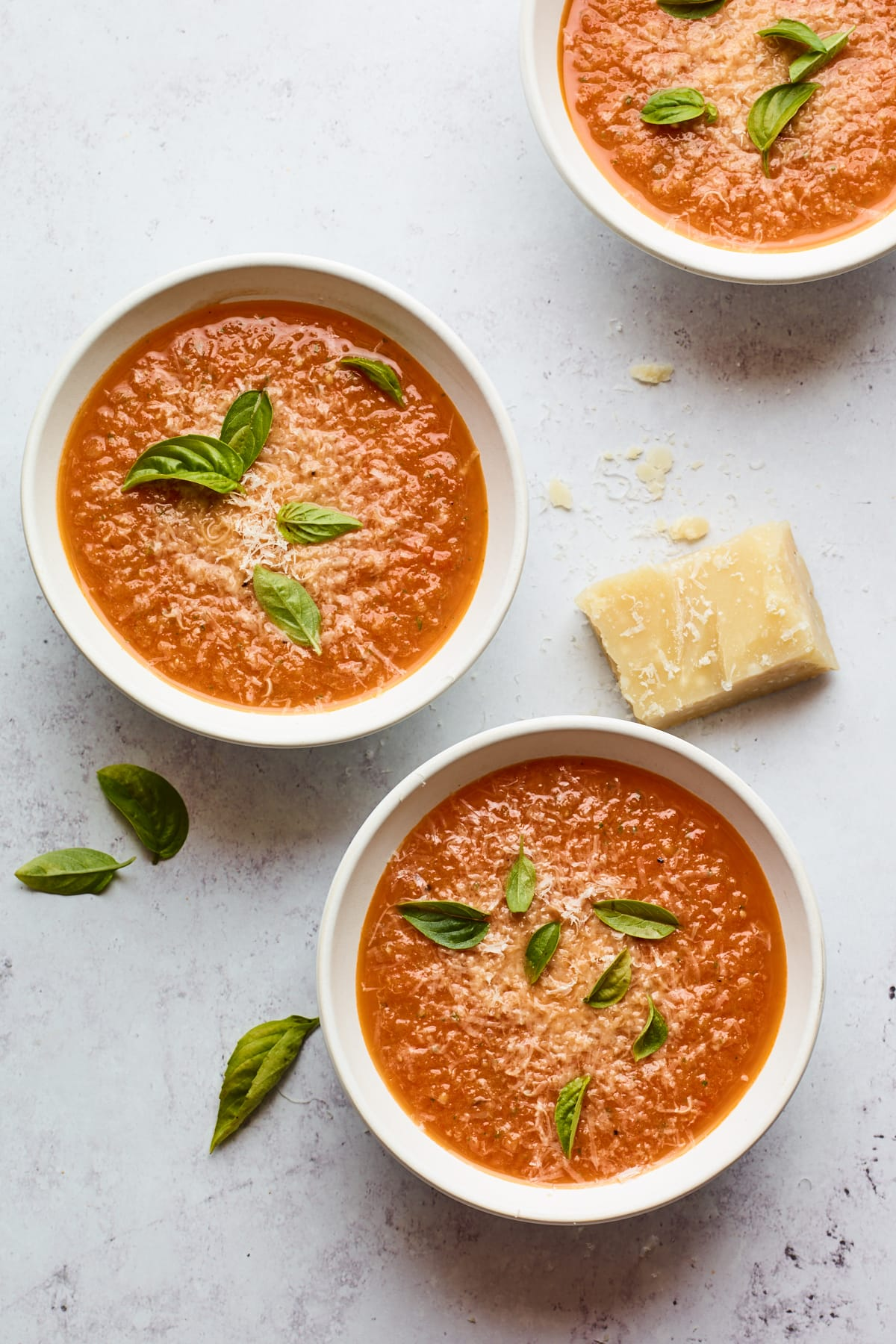 Três taças contendo sopa de manjericão de tomate. Cada tigela é coberta com folhas frescas de manjericão e há uma cunha de queijo parmesão que descansa perto das tigelas.