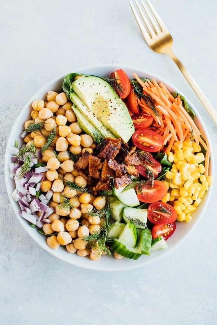 Salada de espiga vegan com bacon tempeh e grão-de-bico numa tigela branca com garfo dourado.